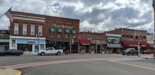 A small town street scene featuring shops, a barber, and an American flag, with cloudy skies above.