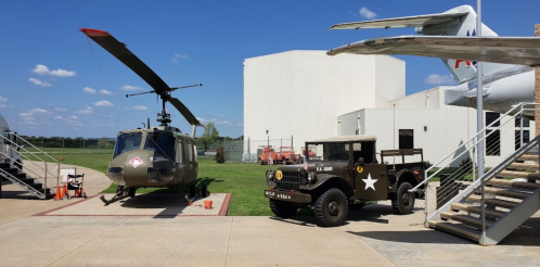 A military helicopter and vintage army vehicle displayed outdoors near a building and aircraft under a clear blue sky.