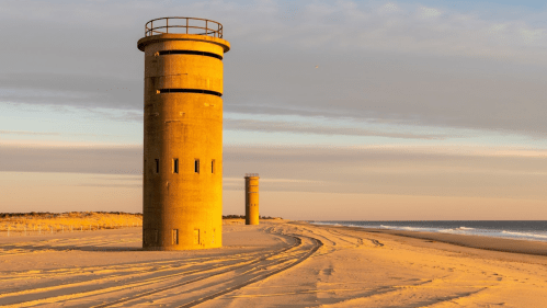 Two tall, cylindrical concrete towers on a sandy beach at sunset, with gentle waves in the background.