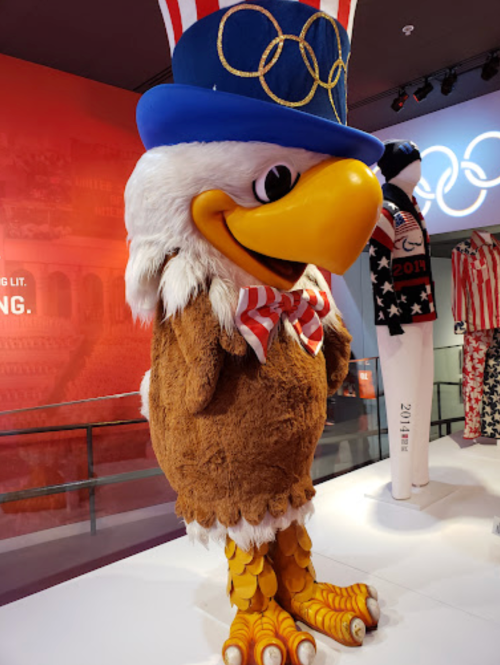 A large, cartoonish eagle mascot wearing a blue top hat and Olympic rings, with patriotic red, white, and blue accents.