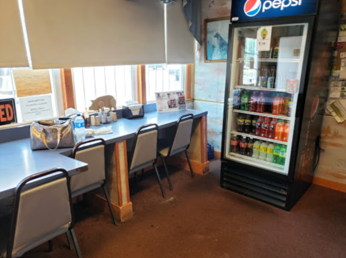 A small dining area with a table, chairs, and a Pepsi vending fridge filled with drinks. Windows let in natural light.