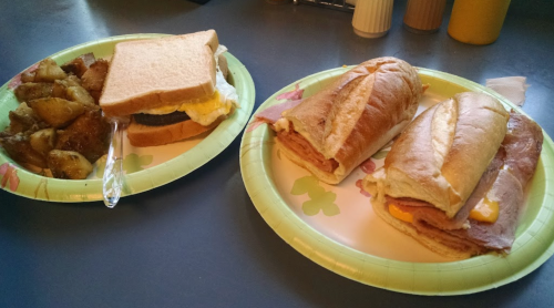 A plate with two sandwiches and a side of seasoned potatoes on a table. One sandwich has egg, the others have ham and cheese.
