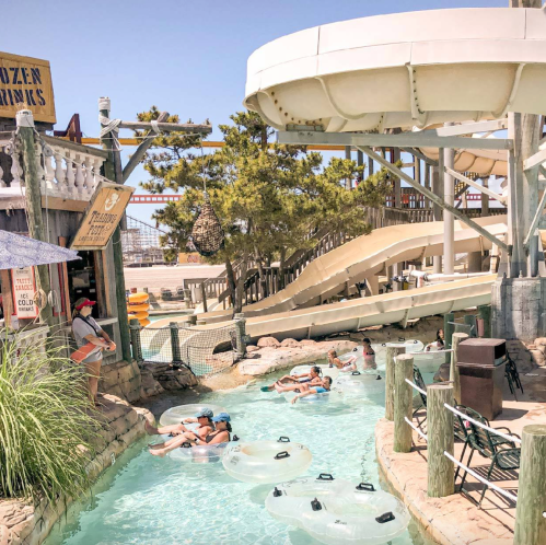 A water park scene with a lazy river, water slides, and people floating on inner tubes under a sunny sky.