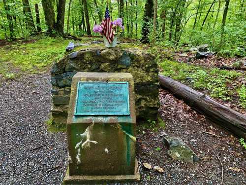 A stone memorial in a forest, featuring a plaque and a small American flag with flowers on top.