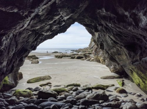 View from inside a rocky cave, looking out at a sandy beach and calm ocean under a cloudy sky.
