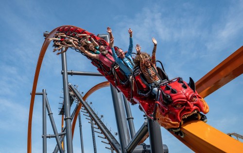 A roller coaster with riders raising their hands, featuring a red dragon design at the front, against a blue sky.