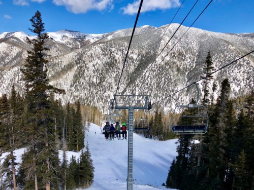 Skiers on a chairlift ascend a snowy mountain, surrounded by tall pine trees and scenic peaks under a blue sky.