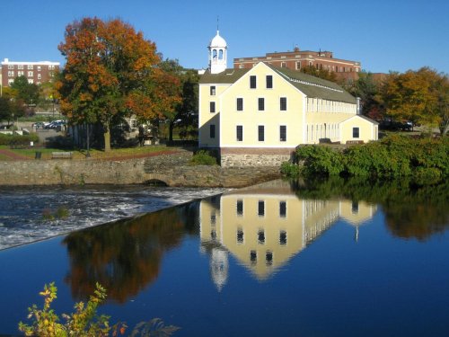 A yellow building with a white steeple reflects in a calm river, surrounded by autumn trees and clear blue skies.