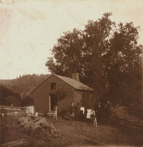 A vintage photo of a small house with a group of people standing outside, surrounded by trees and a rural landscape.