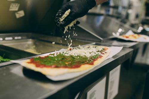 A hand in a black glove sprinkles cheese over a long pizza topped with sauce and herbs on a countertop.