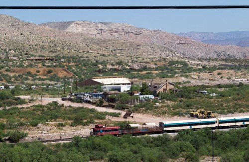 A train moves through a desert landscape with mountains in the background and scattered buildings and vehicles.