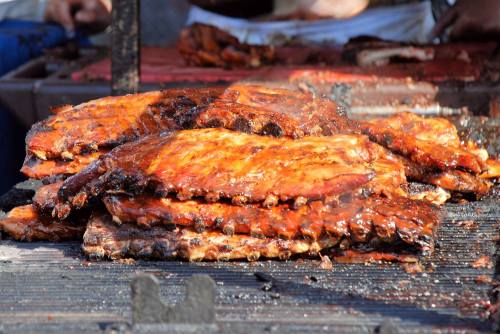 Grilled ribs stacked on a barbecue grill, glistening with sauce and smoke rising in the background.