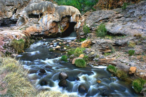 A serene river flows past rocky formations and grassy patches, with a cave entrance visible in the background.