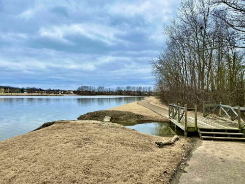 A calm lake scene with a wooden dock, grassy shore, and bare trees under a cloudy sky.