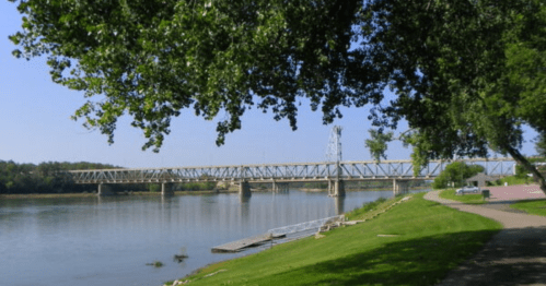 A riverbank scene featuring a bridge, trees, and a pathway along the water under a clear blue sky.