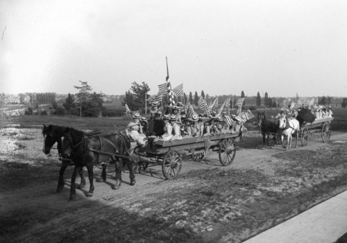 A horse-drawn wagon filled with people waving American flags, traveling along a dirt road in a rural setting.