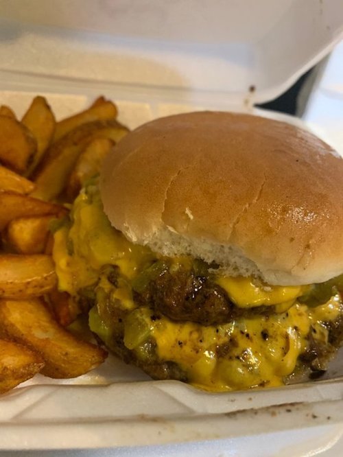 A cheeseburger with melted cheese and pickles, served with crispy potato wedges in a takeout container.