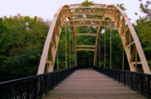 A wooden bridge with an arched metal frame, surrounded by lush green trees.