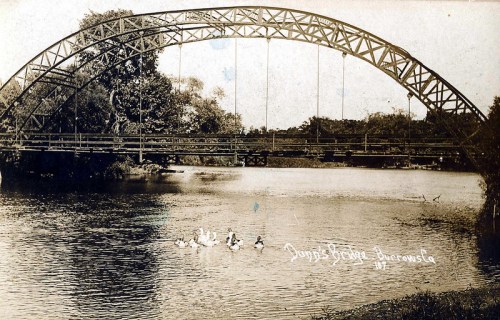 Black and white photo of a bridge over a river, with ducks swimming below and trees in the background.