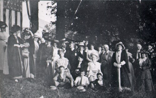 A large group of people in early 20th-century attire poses outdoors near an American flag.
