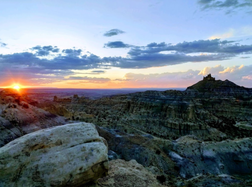 A stunning sunset over rugged terrain, with dramatic clouds and a distant rock formation silhouetted against the sky.