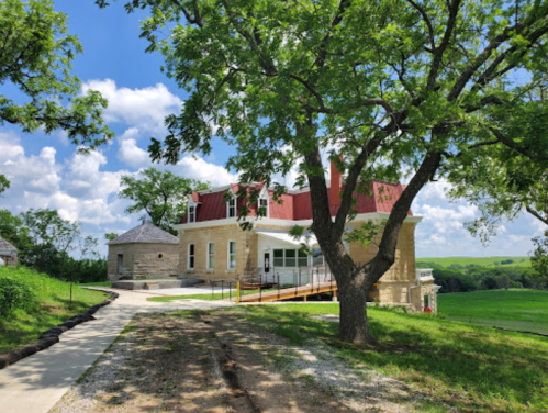 A historic stone building with a red roof, surrounded by trees and green fields under a blue sky with clouds.