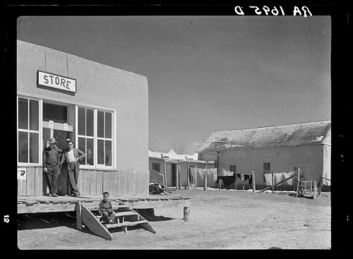 A small store in a desert town, with two men standing outside and a child sitting on the steps. Nearby buildings and laundry visible.