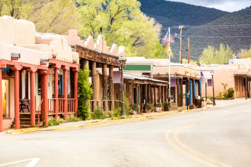 A quiet street lined with adobe-style buildings and colorful storefronts, surrounded by green trees and mountains.