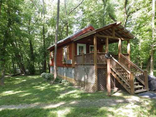 A small red cabin with a porch, surrounded by lush green trees and grass.