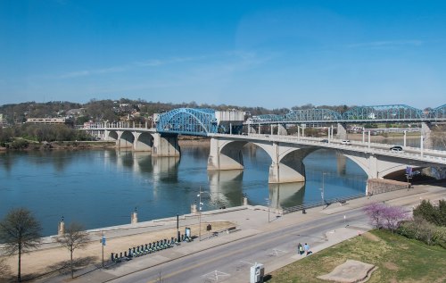 A blue bridge spans a river, with a park and walking path visible along the shore under a clear blue sky.