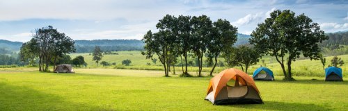 A serene campsite with colorful tents set on green grass, surrounded by trees and rolling hills under a blue sky.