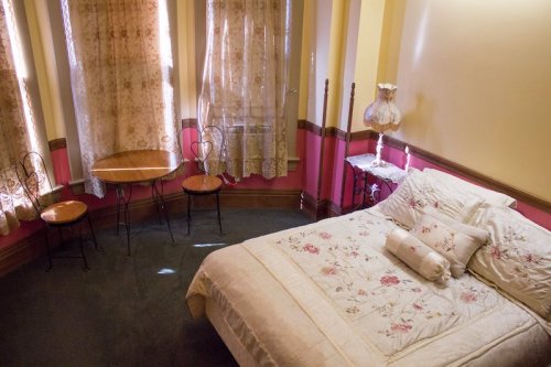Cozy bedroom with a floral bedspread, small table, chairs, and lace curtains letting in soft light.