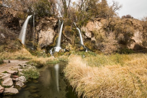 A serene landscape featuring three waterfalls cascading into a calm stream, surrounded by autumn foliage and tall grass.