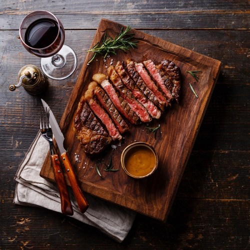 A perfectly cooked steak, sliced and garnished with herbs, served on a wooden board with a side of sauce and a glass of red wine.