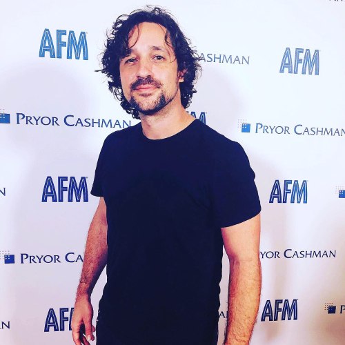 A man with curly hair stands confidently in a black t-shirt against a backdrop featuring "AFM" and "Pryor Cashman."