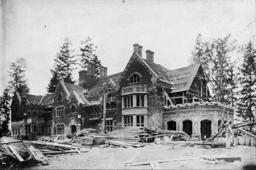 Historic black-and-white photo of a large, partially constructed mansion surrounded by trees and building materials.