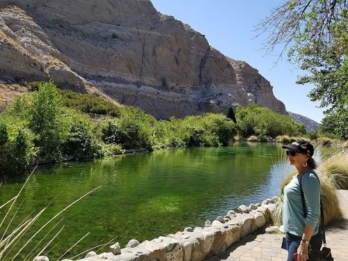 A woman stands by a green river surrounded by lush greenery and rocky cliffs under a clear blue sky.
