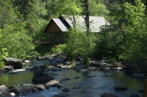 A serene landscape featuring a small cabin by a stream, surrounded by lush greenery and rocky terrain.