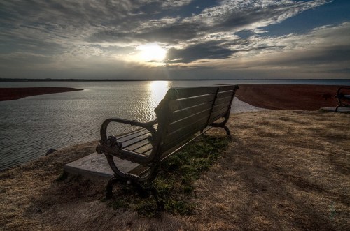 A solitary bench overlooks a calm lake at sunset, with clouds reflecting the warm light.