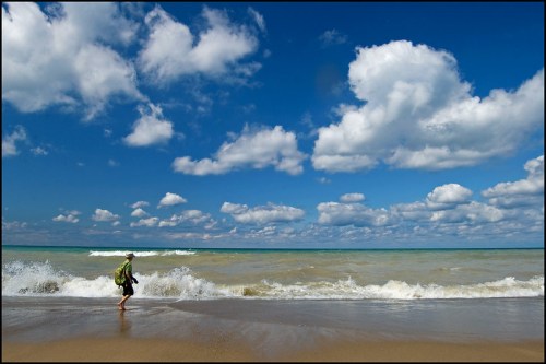 A person walks along a sandy beach, waves crashing at their feet under a bright blue sky with fluffy clouds.