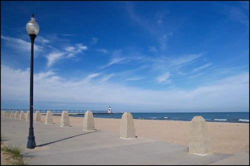 A serene beach scene with a lamp post, concrete barriers, and a distant lighthouse under a blue sky.