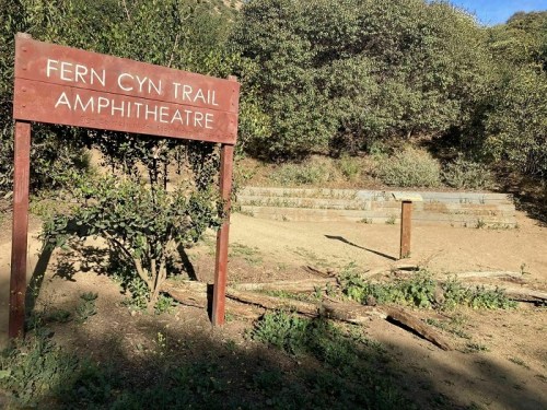 Sign for "Fern Cyn Trail Amphitheatre" in a natural setting with greenery and a dirt path.