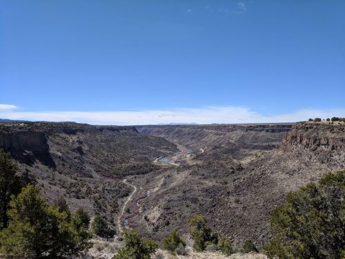 A panoramic view of a deep canyon with a winding river, surrounded by rocky cliffs and sparse vegetation under a clear blue sky.
