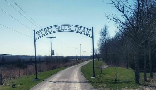 Sign for Flint Hills Trail over a dirt path, flanked by trees and power lines under a clear blue sky.