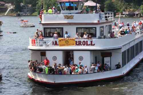 A large boat filled with people cruises on a lake, with banners and flags visible on the deck.