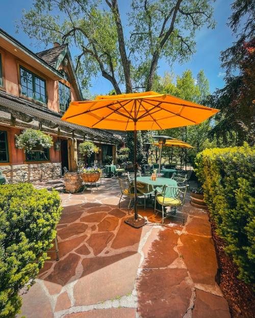 A sunny outdoor patio with orange umbrellas, green tables, and lush greenery surrounding a charming stone house.