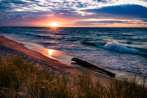 Sunset over a beach with gentle waves, sandy shore, and grass in the foreground, creating a serene coastal scene.