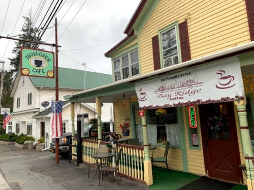 A quaint café with a yellow exterior, outdoor seating, and a sign reading "We Proudly Serve Bay Ridge Coffee."