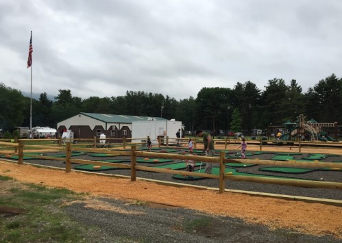 A playground with mini-golf and children playing, surrounded by trees and a cloudy sky. An American flag is visible.