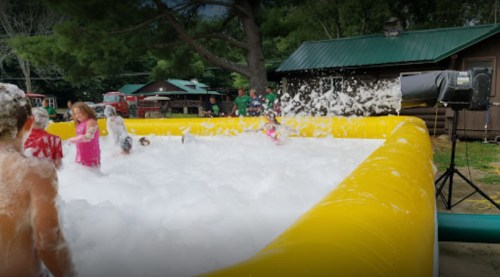Children playing in a large inflatable pool filled with foam, surrounded by trees and a cabin in the background.